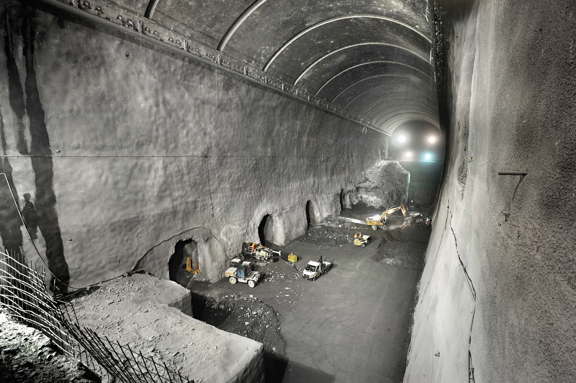Das Bild zeigt eine typische Szene aus dem Bau eines Kraftwerks. Der Tunnel ist ein langer, dunkler und feuchter Raum. Die Wände des Tunnels sind aus Beton und werden beleuchtet. Am Boden des Tunnels liegen Schienen, auf denen der Bagger fährt.
